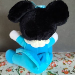 May include: A plush toy of a cartoon mouse wearing a blue outfit. The mouse has black ears and a black nose. The toy is sitting on a patterned fabric.