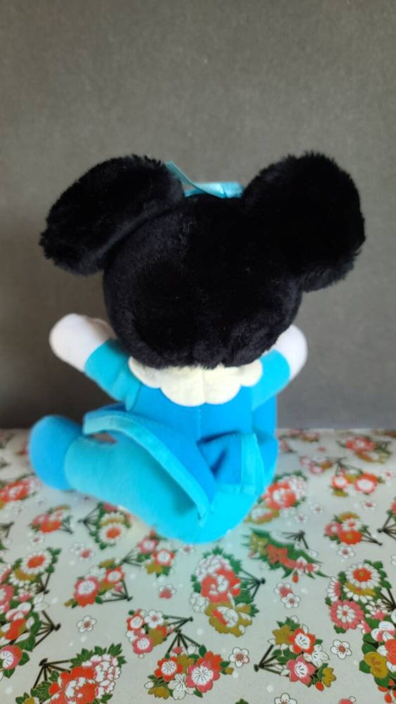 May include: A plush toy of a cartoon mouse wearing a blue outfit. The mouse has black ears and a black nose. The toy is sitting on a patterned fabric.
