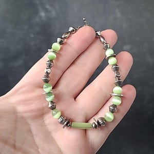 May include: A beaded bracelet featuring light green cat's eye beads and silver-toned metal accents. The bracelet has a mix of round and geometric beads, with a central rectangular bead. The clasp is a simple chain.