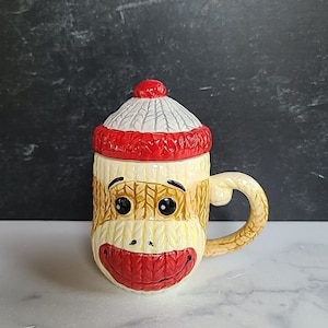 Peut inclure: Une tasse à café en céramique en forme de singe chaussette, avec un couvercle en forme de bonnet rouge et blanc. La tasse a un corps jaune, une bouche rouge et des yeux noirs, et une anse incurvée. La tasse est sur une surface blanche.