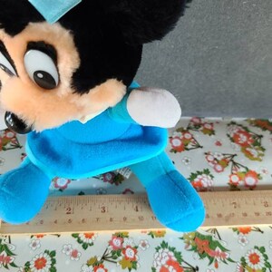 May include: A blue and black plush toy mouse wearing a blue dress and a blue bow. The toy is approximately 10 inches tall and is sitting on a white and floral patterned surface.