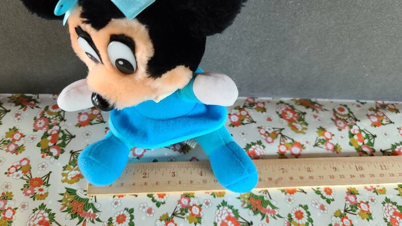 May include: A blue and black plush toy mouse wearing a blue dress and a blue bow. The toy is approximately 10 inches tall and is sitting on a white and floral patterned surface.