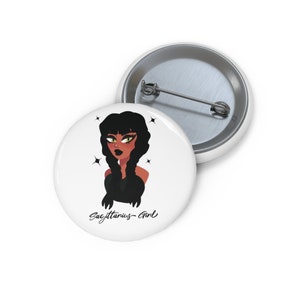 May include: A white button with a cartoon illustration of a woman with long black hair and dark brown skin. She is wearing a black top and has green eyes. The text "Sagittarius Girl" is written below the illustration.