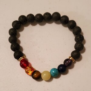 Puede incluir: Un brazalete de roca de lava negra con detalles dorados y cuentas de piedras preciosas de colores. Las cuentas son rojas, naranjas, amarillas, verdes, azules y moradas.