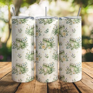 May include: Three white insulated tumblers with a floral and frog design. Each tumbler features a repeating pattern of green frogs wearing flower crowns, surrounded by white and yellow flowers, and green leaves. The tumblers have silver straws.