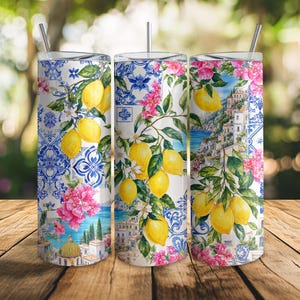 Può includere: Un bicchiere cilindrico in acciaio inossidabile con cannuccia, caratterizzato da una vivace stampa di ispirazione mediterranea con limoni gialli, foglie verdi, fiori di bouganville rosa, piastrelle blu e bianche e un villaggio costiero.