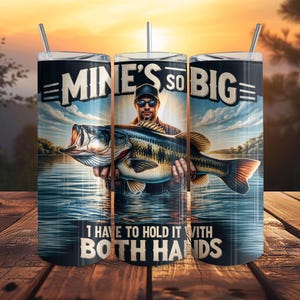 May include: A stainless steel tumbler with a fishing theme. The tumbler features a man holding a large fish with the text "MINE'S SO BIG I HAVE TO HOLD IT WITH BOTH HANDS". The background shows a lake and sunset.