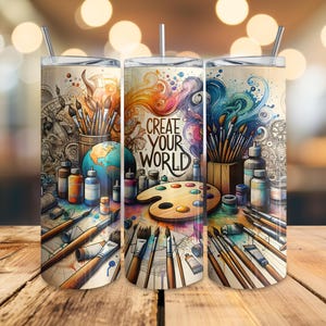 May include: Three stainless steel tumblers with lids and straws, featuring a colorful artistic design. The design includes paintbrushes, paint bottles, a globe, and the text "Create Your World" on a light beige background. The tumblers are set on a wooden surface.