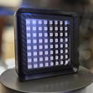 May include: A square electronic display device featuring a grid of small, illuminated white LED lights arranged in rows and columns. The display is encased in a black frame with a textured carbon fiber pattern on the front border surface.