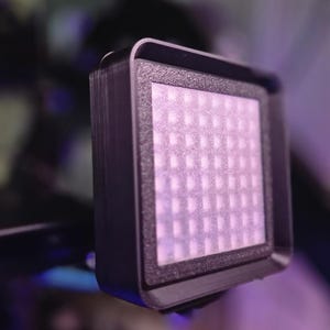 May include: A small, square LED light panel with a grid of small lights. The panel is encased in a black frame and is mounted on a black support structure. The lights are emitting a soft, diffused light.