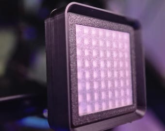 May include: A small, square LED light panel with a grid of small lights. The panel is encased in a black frame and is mounted on a black support structure. The lights are emitting a soft, diffused light.