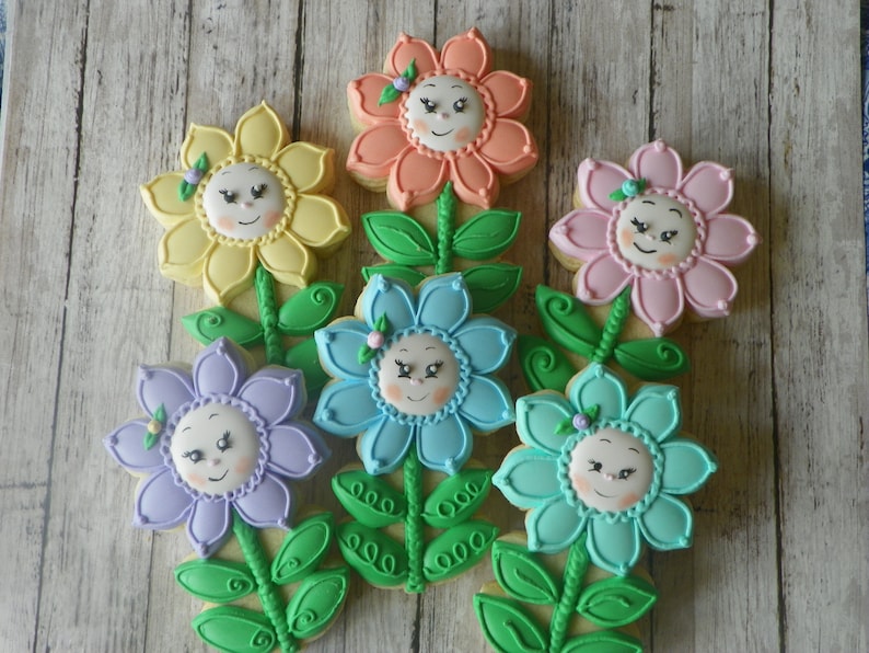 May include: Six decorated sugar cookies shaped like flowers with smiling faces. The cookies are in various colours, including yellow, orange, pink, purple, blue, and green.