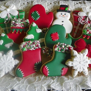 May include: A collection of Christmas-themed cookies in various shapes, including mittens, stockings, Christmas trees, snowflakes, and gift boxes. The cookies are decorated with red, green, and white icing, and sprinkles. The cookies are arranged on a white lace surface.
