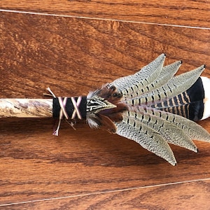 May include: A ceremonial feather fan featuring a polished wooden handle with a marbled brown pattern. The base of the feathers is wrapped in black leather with a decorative white and purple crisscross lacing, accented by brown and grey patterned feathers.