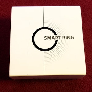 Könnte beinhalten: Eine weiße Schachtel mit den Worten "SMART RING" in schwarzer Schrift. Die Schachtel hat ein schwarzes kreisförmiges Design mit konzentrischen Kreisen. Die Schachtel befindet sich auf einer roten Oberfläche.