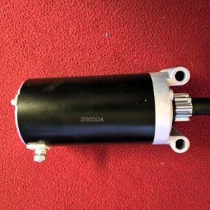 Peut inclure: Un démarreur cylindrique noir avec des supports de montage argentés et un engrenage à une extrémité. Le numéro "250304" est imprimé sur le corps noir. Le moteur est posé sur un fond rouge.