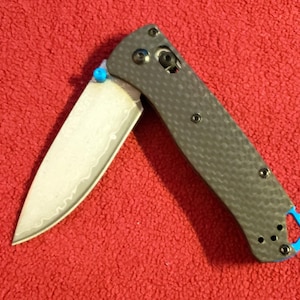 Könnte beinhalten: Ein Klappmesser mit gemusterter Klinge und grauem Griff. Die Klinge ist teilweise geöffnet und zeigt ein detailliertes Design. Der Griff hat eine strukturierte Optik und blaue Akzente. Das Messer liegt vor rotem Hintergrund.