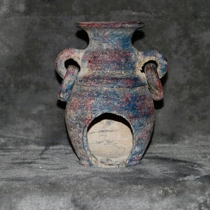 May include: A textured ceramic vessel featuring a mottled blue and reddish-brown finish. The piece has a wide neck, two side handles with circular openings, and a large, arched cutout at the base, revealing a light-colored interior surface.