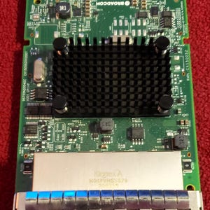 Op de afbeelding: Een groene printplaat met diverse elektronische componenten, waaronder chips, condensatoren en een koellichaam. De printplaat heeft een zilveren metalen plaat met de tekst "Kinnex A" en een reeks blauwe lampjes aan de onderkant. De printplaat ligt op een rode ondergrond.