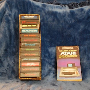 May include: A vertical stack of twelve Atari video game cartridges housed in a rectangular woven basket, positioned next to a paperback book titled The User's Guide to Atari 400 800 Video Computers, Software, & Peripherals by Consumer Guide.