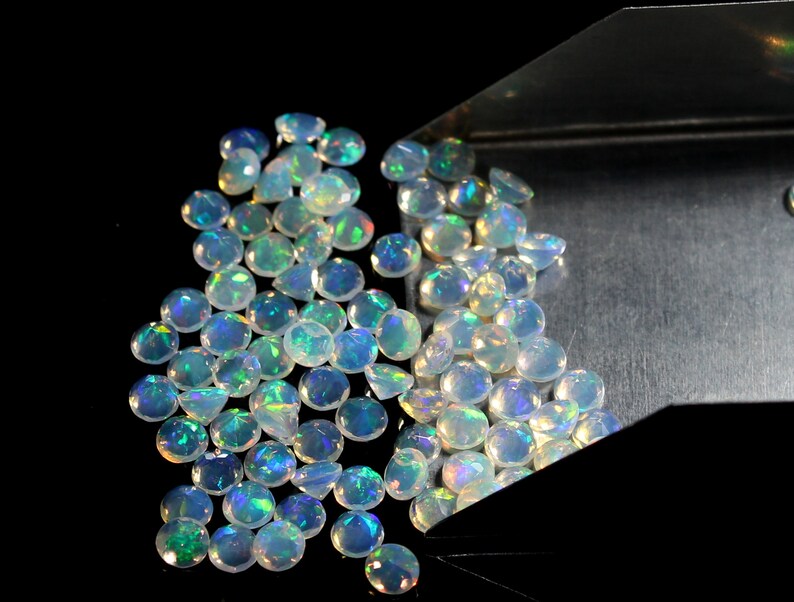 Pode incluir: Uma cole&ccedil;&atilde;o de opalas facetadas, exibindo uma gama de cores iridescentes, incluindo azul, verde e amarelo. As pedras preciosas s&atilde;o redondas e est&atilde;o espalhadas, algumas derramando de uma bandeja de metal contra um fundo preto.