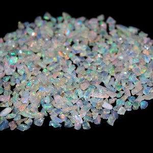 May include: A pile of small, iridescent opal chips. The chips are mostly white with flashes of blue, green, and orange. The gemstones are irregularly shaped and reflect light, creating a sparkling effect against the black background.