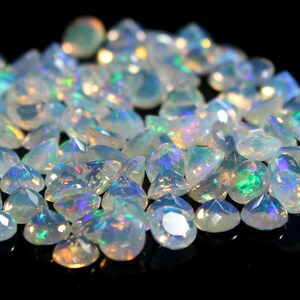 Pode incluir: Uma cole&ccedil;&atilde;o de pedras preciosas de opala facetadas, exibindo uma variedade de formas e tamanhos. As opalas t&ecirc;m uma cor de base branca leitosa com flashes iridescentes de verde, azul e amarelo. As pedras preciosas est&atilde;o espalhadas em uma superf&iacute;cie preta.