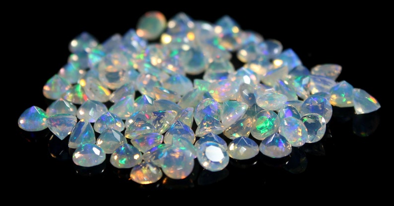 Pode incluir: Uma cole&ccedil;&atilde;o de pedras preciosas de opala facetadas, exibindo uma variedade de formas e tamanhos. As opalas t&ecirc;m uma cor de base branca leitosa com flashes iridescentes de verde, azul e amarelo. As pedras preciosas est&atilde;o espalhadas em uma superf&iacute;cie preta.