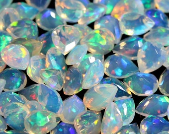 Pode incluir: Uma coleção de pedras preciosas de opala facetadas e iridescentes. As opalas têm várias formas, incluindo lágrimas e ovais, e exibem uma gama de cores, incluindo azul, verde e amarelo. As pedras preciosas estão espalhadas sobre uma superfície escura.