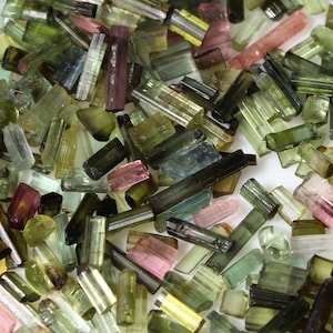 Op de afbeelding: Een verzameling kleine, kleurrijke en doorschijnende toermalijnkristallen. De kristallen zijn in verschillende tinten groen, roze en blauw. Ze zijn allemaal langwerpig en hebben een glad, gepolijst oppervlak.