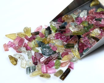 May include: A collection of colorful, raw, rough, and tumbled gemstones. The gemstones are various shades of pink, green, blue, and yellow. The gemstones are in a metal scoop.