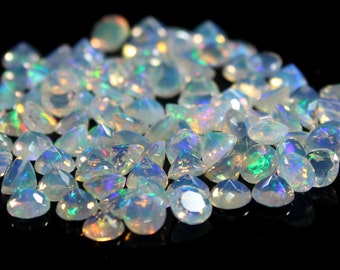May include: A collection of small, faceted, translucent white opals with iridescent flashes of green, blue, and orange light. Each gemstone is cut into a triangular shape with multiple facets that reflect light against a dark background.