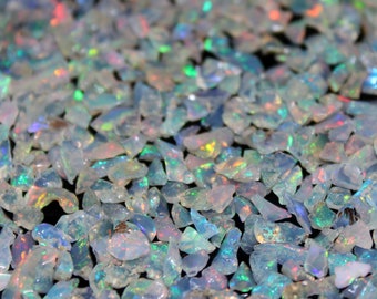 May include: A close-up view of a collection of small, irregularly shaped opal gemstones. The opals are primarily white and translucent, with flashes of iridescent color including green, blue, and red. The gemstones are scattered across the surface.