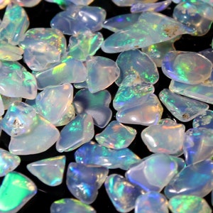 Könnte beinhalten: Eine Sammlung polierter Opal-Edelsteine. Die Opale sind überwiegend weiß mit schillernden Blitzen in Grün, Blau und Gelb. Die Steine variieren in Größe und Form und haben eine glatte, glänzende Textur. Der Hintergrund ist schwarz.