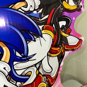 May include: A close-up of a cardboard cutout of Sonic the Hedgehog and Shadow the Hedgehog. Sonic is blue with white gloves and red shoes. Shadow is black with red eyes and red shoes. The background is pink with white stars.