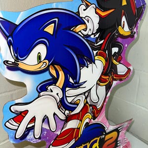 May include: A cardboard cutout of Sonic the Hedgehog and Shadow the Hedgehog from the video game Sonic Adventure 2. The characters are posed in a dynamic action pose. The background is a purple and blue gradient with stars. The text "Sonic Adventure 2" and "SEGA Dreamcast" is visible at the bottom of the cutout.