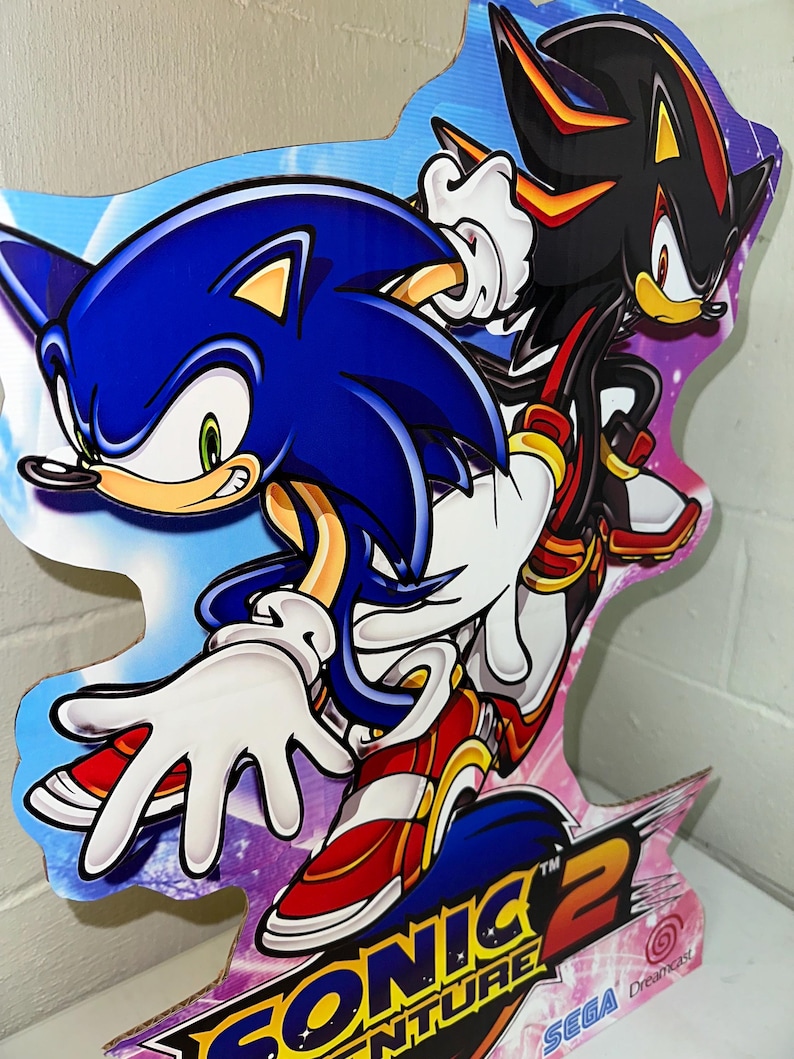 May include: A cardboard cutout of Sonic the Hedgehog and Shadow the Hedgehog from the video game Sonic Adventure 2. The characters are posed in a dynamic action pose. The background is a purple and blue gradient with stars. The text "Sonic Adventure 2" and "SEGA Dreamcast" is visible at the bottom of the cutout.