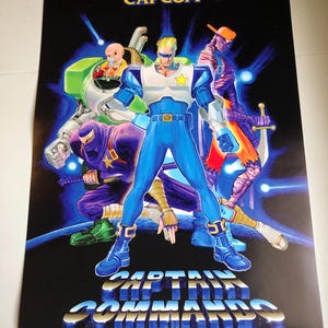 May include: A CAPCOM Captain Commando poster. The image shows a central character in a blue suit with a star emblem, surrounded by other costumed characters. The background is black with blue and white accents.