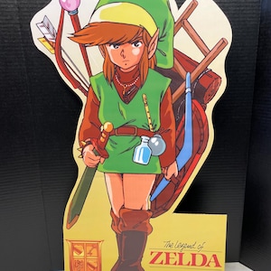 Puede incluir: Un recorte de cartón de Link del videojuego The Legend of Zelda. Link lleva una túnica verde, pantalones marrones y un sombrero verde. Lleva una espada, un escudo y un arco y flechas. El recorte está sobre un fondo amarillo con las palabras "The Legend of Zelda" y "Nintendo" impresas en él.