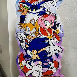 May include: A cardboard cutout of Sonic the Hedgehog and his friends from the video game Sonic Adventure. The characters are arranged in a stack, with Sonic at the bottom, followed by Tails, Amy, Knuckles, and Shadow. The cutout is printed with a purple background and the text "Sonic Adventure" in large, white letters. The bottom of the cutout features the Sega Dreamcast logo and the text "Sonic Team".