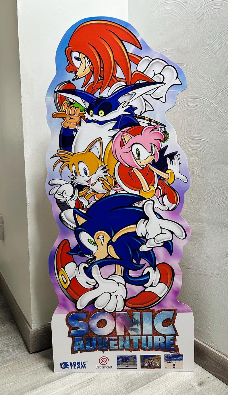 May include: A cardboard cutout of Sonic the Hedgehog and his friends from the video game Sonic Adventure. The characters are arranged in a stack, with Sonic at the bottom, followed by Tails, Amy, Knuckles, and Shadow. The cutout is printed with a purple background and the text "Sonic Adventure" in large, white letters. The bottom of the cutout features the Sega Dreamcast logo and the text "Sonic Team".