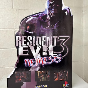 May include: A cardboard cutout of a zombie-like figure with a large mouth and a grotesque face. The figure is holding a large, black pipe. The text "RESIDENT EVIL 3 NEMESIS" is printed in white on a black background. The text "CAPCOM" and "PlayStation" are also printed on the cutout.
