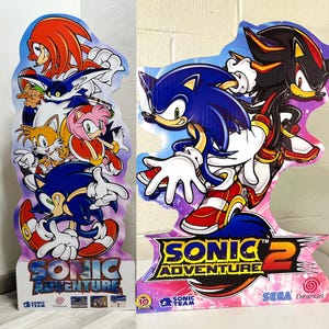May include: A cardboard cutout of Sonic the Hedgehog characters from the video game Sonic Adventure 2. The characters are Sonic, Shadow, Knuckles, Tails, Amy, and Big. The cutout is on a pink and purple background with the game's logo and title. The text "Sonic Adventure 2" is in a yellow, red, and blue font. The text "SEGA Dreamcast" is in a white font.