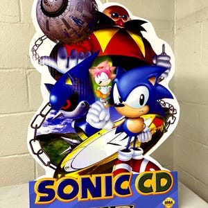 Könnte beinhalten: Ein Papp-Ausschnitt von Sonic the Hedgehog, Tails, Knuckles und Dr. Robotnik aus dem Sega Genesis-Spiel Sonic CD. Die Figuren stehen vor einem blauen Himmel mit Wolken. Der Ausschnitt befindet sich auf einer weißen Oberfläche. Der Text "Sonic CD" ist in blauen und grünen Buchstaben auf gelbem Hintergrund gedruckt. Das Sega-Logo ist in blauen und weißen Buchstaben auf blauem Hintergrund gedruckt.