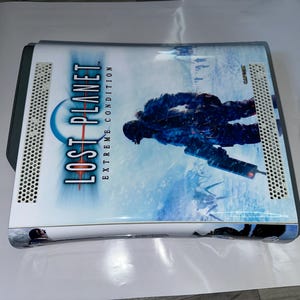 Pode incluir: Um console de jogos branco e cinza com o jogo "Lost Planet: Extreme Condition" impresso na parte superior. A capa do jogo apresenta uma paisagem gelada azul e branca com uma figura em um traje de inverno.