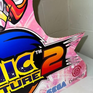 May include: A pink and blue cardboard cutout of Sonic Adventure 2 for the Sega Dreamcast. The cutout features the game's logo and the Sega and Dreamcast logos.