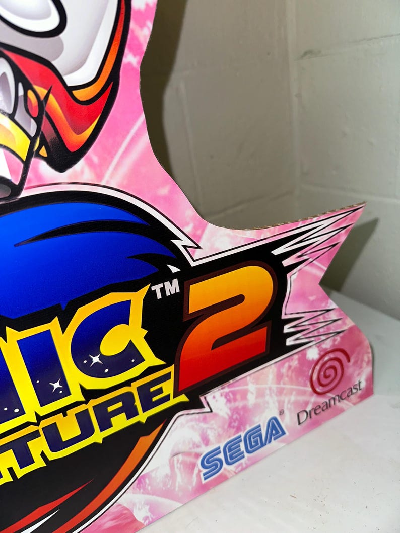 May include: A pink and blue cardboard cutout of Sonic Adventure 2 for the Sega Dreamcast. The cutout features the game's logo and the Sega and Dreamcast logos.