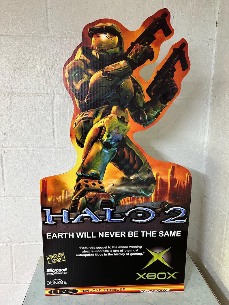 May include: A cardboard cutout of a green and blue video game character holding two guns. The character is standing in front of a cityscape with a fiery orange background. The text "HALO 2" is in large white letters at the top of the cutout. The text "EARTH WILL NEVER BE THE SAME" is in white letters below the title. The text "ONLY ON XBOX" is in a white box with a black border. The text "Microsoft game studios" is in white letters with a black border. The text "BUNGIE" is in white letters with a black border. The text "LIVE ONLINE ENABLED" is in white letters with a black border. The text "www.xbox.com" is in white letters with a black border. The Xbox logo is in green and white.