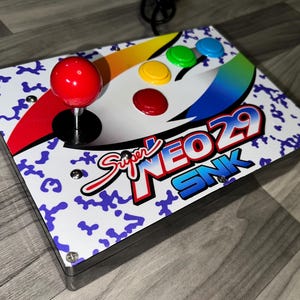 Può includere: Un controller arcade rettangolare con una superficie bianca con motivo mimetico blu, un joystick rosso e quattro pulsanti colorati in giallo, verde, blu e rosso. Il controller mostra il testo Super NEO 29 SNK in un carattere in grassetto.