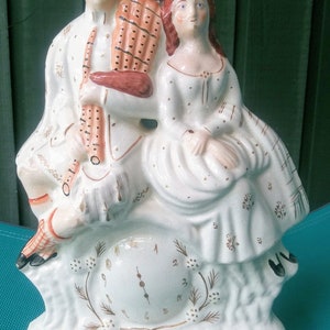 Könnte beinhalten: Eine weiße Keramikfigur eines Paares, ein Mann mit Hut und eine Frau in einem Kleid, die auf einer Uhr sitzen. Der Mann hält eine Dudelsackpfeife und die Frau hält ihre Hände in ihrem Schoß. Das Zifferblatt der Uhr hat römische Ziffern und die Zeiger zeigen auf 12 und 3.